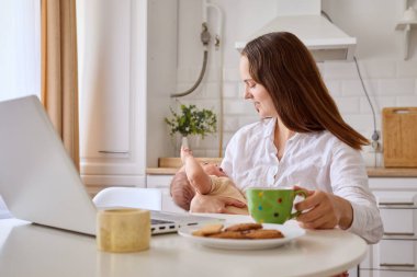 Bir anne, bebeğini tek koluyla tutarken iş ve ebeveynliği dengeler, masada dizüstü bilgisayar ve bir tabak kurabiye ile. Mutfak aydınlık ve davetkar..