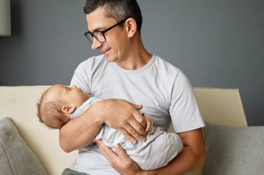 Bir baba sevgiyle yeni doğmuş bebeğini modern bir oturma odasına yatırır. İkisi de sakin ve bağlı görünürler. Güneş ışığı onların hassas etkileşimlerini vurgular.