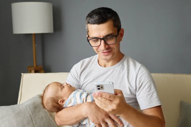 Bir baba bebeğini bir koluyla tutarken diğer koluyla akıllı telefonunu kullanır. İyi aydınlatılmış bir oturma odasında, huzurlu bir atmosfer yaratarak rahatça oturuyorlar..
