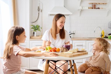 Bir anne, güneşli bir mutfak masasında iki kızıyla keyifli bir kahvaltı yapar. Sıcak bir atmosferde taze meyve, krep ve kahkahaların tadını çıkarır..