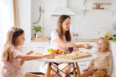 Bir anne kahvaltı masasında kızlarıyla iyi vakit geçirmekten hoşlanır. Aile, sıcak ve sevgi dolu bir atmosfer oluşturan meyve ve tostlardan oluşan bir yemeği paylaşıyor..