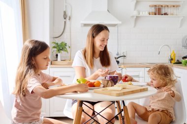 Bir anne, güneşli bir mutfakta iki çocuğuyla birlikte yuvarlak bir masada oturur, kahvaltısını paylaşır ve taze meyvelerle çevriliyken sohbet eder..
