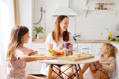 İki çocuk ve bir yetişkin, doğal ışıkla dolu bir mutfakta keyifli bir kahvaltı yaparlar. Yuvarlak bir masada taze meyve ve hamur işleriyle otururlar ve sıcak bir atmosfer yaratırlar..