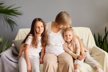 Two young girls sit beside an adult, sharing smiles and laughter in a comfortable living room. The atmosphere is warm, filled with affection and contentment, surrounded by greenery. clipart