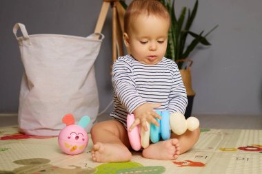 Bir bebek, neşeli bir paspasın üzerinde bağdaş kurarak, yumuşak, renkli oyuncak yüzüklerle kendini eğlendirir. Odada doğal dekor ve yumuşak ışık var. Sıcak bir atmosfer yaratıyor..