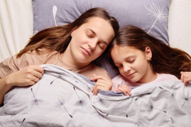 Bir anne ve küçük kızı rahat bir yatak odasında yan yana mışıl mışıl uyuyor. Yumuşak bir battaniyeyle kaplanmışlar, sıcaklık ve sükunet yayıyorlar..