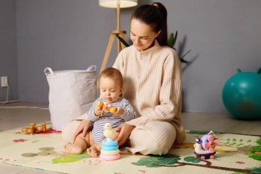 Bir anne bebeğiyle yerde oturur renkli oyuncaklar ve yumuşak bir paspasın arasında oyun zamanının tadını çıkarır. Rahatlamış atmosferde bir lamba ve neşeli bir dekor var..