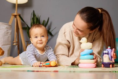 Neşeli bir bebek yumuşak bir mindere uzanıp renkli oyuncaklarla oynayarak annelerinin onlarla etkileşime geçmesini ve rahat, parlak bir yaşam alanında keşif ve gelişmeyi teşvik eder..
