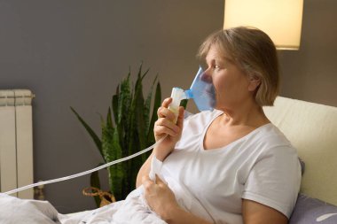 Nebulizör kullanarak yatakta oturan bir kadın solunum sorununu ele alıyor. Odaklanmış ve sakin görünüyor, evde bakımın sağlıkla ilgilenmedeki önemini vurguluyor..