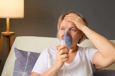 Yaşlı bir kadın rahat oturma odasında bir kanepede oturuyor, solunum tedavisi için nebulizör kullanıyor. Alnını tutarken endişeli görünüyor, bir lamba hafifçe mekanı aydınlatıyor..