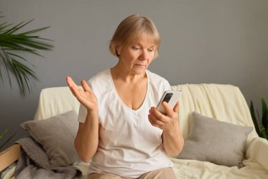 Yaşlı bir kadın rahat bir kanepede oturuyor, akıllı telefonunu dalgın bir ifadeyle tutuyor. Etrafı yumuşak yastıklarla çevrili, önemli bir konuşma veya görevle meşgul gibi görünüyor..