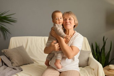 Neşeli bir kadın, bitkilerle ve rahat dekorlarla dolu modern bir oturma odasında bir bebeği kucaklıyor. Atmosfer sıcak ve davetkar, bağlarını vurguluyor..
