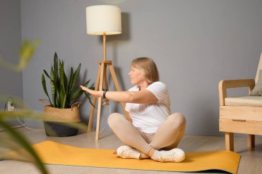 Bir kadın yoga minderinde oturmuş, iyi aydınlatılmış bir odada yoga yapıyor. Etrafı kapalı bitkilerle ve rahat mobilyalarla çevrili. Huzurlu bir atmosfer yaratıyor..