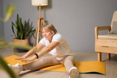 Altmışlı yaşlarda bir kadın, sarı yoga minderinin üzerinde esneme egzersizleri yapıyor. Rahat, aydınlık bir oturma odasında. Yeşil bitkiler dekora ferahlatıcı bir dokunuş ekler..