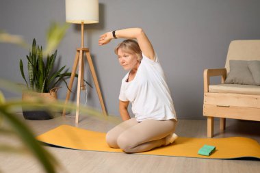 Sakin bir ortamda yaşlı bir kadın sarı bir minderde yoga yapıyor. Bitkiler ve basit mobilyalarla çevrili bir şekilde yerde otururken kolunu uzatıyor..