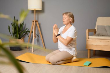 Altmışlı yaşlarda bir kadın evdeki sarı bir hasırda yoga yapıyor ve dua pozisyonunda ellerini kullanarak farkındalık egzersizi yapıyor. Sıcak atmosferde bitkiler ve rahat bir sandalye var..