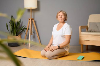 Son sınıf öğrencisi bir kadın yoga minderinde oturuyor, meditasyonla meşgul, iyi ışıklandırılmış bir oturma odasında bitkiler ve rahat mobilyalarla. Ortam sakin ve davetkar..