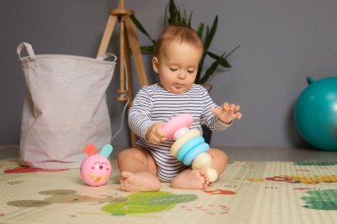 Küçük bir çocuk canlı oyuncaklarla dolu bir oyun hasırının üzerinde oturuyor. Neşeli bir dekorla çevrili oda sıcak ve davetkar bir atmosfere sahiptir. Bebek istiflemeyi ve keşfetmeyi seviyor..