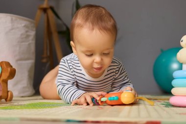 Küçük bir çocuk güneşli bir odada yumuşak bir paspasın üzerinde yatarken parlak, renkli bir oyuncağa odaklanır. Atmosfer neşeli ve eğlenceli. Keşif için mükemmel..