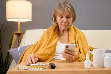 A senior woman wrapped in a cozy blanket sits on a couch, focused on her phone. Nearby, various medications and a warm drink are visible, hinting at her health condition. clipart