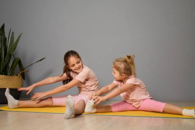 İki genç kız, parlak bir odada sarı bir hasırın üzerinde esneme egzersizleri yapıyorlar. Yoga çalışmaları sırasında gülümserler ve birbirlerini cesaretlendirirler..