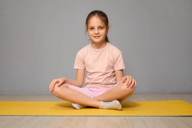 Genç bir kız, sarı yoga minderinin üzerinde bağdaş kurarak oturuyor. Hafif pembe bir gömlek ve pembe bir şort giyerek, yoga pratiği yaparak hafifçe gülümsüyor..