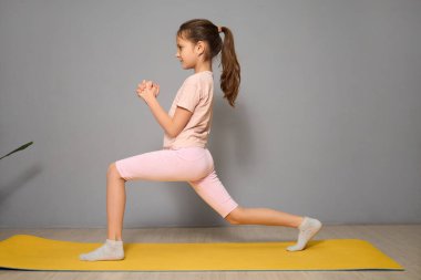 Genç bir kız sarı yoga minderinin üzerinde hamlesini yapıyor. Odaklanmış ve az dekorlu parlak bir odaya yerleştirilmiş. Bu da bir eğitim seansına işaret ediyor..