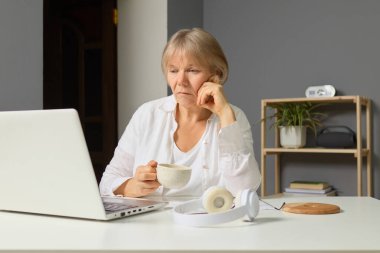 Yaşlı bir kadın, elinde bir fincan kahve tutarken dizüstü bilgisayarına odaklanmış modern bir odada masa başında oturuyor. Kulaklıklar onun yanında, ve bir bitki alanı süslüyor..