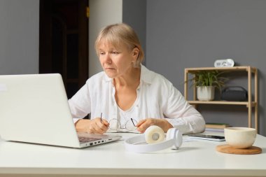 Kısa saçlı son sınıf öğrencisi bir kadın dizüstü bilgisayarına odaklanmış, gözlüklerini ayarlıyor. İşine dalmış, kulaklıkları ve bitkileriyle bir masada oturuyor..