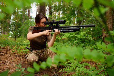 Bir nişancı eğrelti otları ve ağaçlar arasında alçak bir konumda konumlandırılır ve ateş etmeye hazırlanan bir dürbünle dikkatlice odaklanır. Çevre yeşillik ve doğal ışık bakımından zengindir..