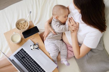 Bir anne yatakta otururken bebeğini emzirir. Önünde bir dizüstü bilgisayar, yanında mısır gevreği ve içecek var. Ev ortamında rahat bir iş ortamı yaratıyor..