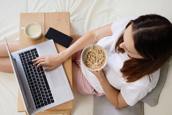 Женщина удобно сидит дома, печатая на ноутбуке с миской овсянки в одной руке и чашкой кофе поблизости, наслаждаясь продуктивным утром.
