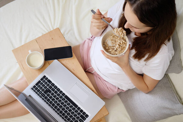 Женщина удобно сидит на кровати и ест миску овсянки. Она балансирует свой ноутбук на коленях и пьет из чашки, начиная свой день с продуктивной утренней рутины.