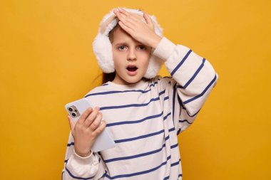 Beyaz kulaklık ve çizgili süveter giyen genç bir kız, elinde akıllı bir telefonla parlak sarı bir arkaplan önünde dururken şaşırmış bir ifade sergiliyor..