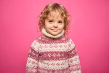 Kıvırcık sarı saçlı neşeli bir çocuk renkli bir kazak giyerken tatlı tatlı gülümsüyor. Parlak pembe arka plan, çocuğun neşeli ifadesini vurguluyor..