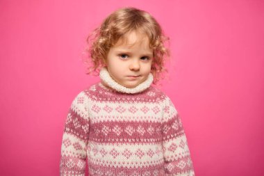 Kıvırcık sarı saçlı ve ciddi bir ifadeye sahip genç bir çocuk parlak pembe bir arka plana karşı duruyor. Örgü örmüş, desenli bir süveter giymişti..