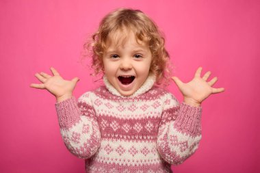 Kıvırcık sarı saçlı genç bir çocuk iki elini de kaldırarak heyecan ve neşe ifade eder. O rahat bir kazak giyer ve canlı pembe bir arka planın önünde durur..