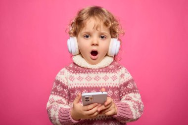 Kıvırcık saçlı bir çocuk beyaz kulaklık takıyor ve akıllı telefon tutarken şaşırıyor. Parlak pembe arka plan neşeli atmosfere ekleniyor..