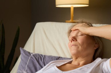 A woman in her seventies sits on a couch with her eyes closed, holding her forehead as she battles fatigue in a comfortable living room filled with soft light. clipart