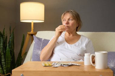 Bir kadın sakin bir yerde oturur, sıcak bir içeceğin tadını çıkarırken ilaçlarını alır. Işıklandırma yumuşak, sabah rutinleri için sakin bir atmosfer yaratıyor..