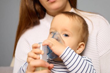 Küçük bir çocuk ebeveyninin kucağında otururken bir astım spreyi tutuyor. Anne, hafif aydınlatılmış bir odada, tıbbi bir anda bakım göstererek rahatlık sağlar..