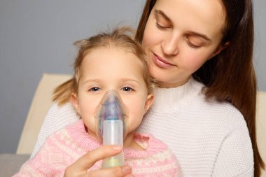 Bir anne, akşam sıcak ve davetkar bir ortamda genç kızına nebulizör kullanmasında yardımcı olur. Çocuk sakin ve tedavisine odaklanmış görünüyor..