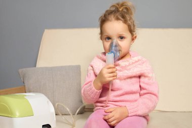 Genç bir kız rahat bir oturma odasında kanepede oturuyor, solunum tedavisi için nebulizör kullanıyor. Yumuşak renkler ve rahat bir ortam sıcak bir atmosfer yaratır..