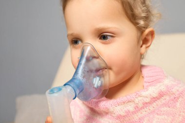 Küçük bir çocuk evde rahat bir şekilde otururken solunum tedavisi için nebulizör kullanıyor. Ortam sıcak ve davetkar, şefkatli bir atmosferi vurguluyor..