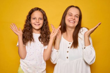 İki dişi, bir anne ve kız, neşeli bir şekilde poz veriyorlar, yüzlerinde gülücüklerle canlı sarı bir zemin üzerinde, ifadelerinde mutluluk ve neşe yayıyorlar..