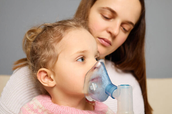 Молодая девушка получает респираторное лечение через туманность, сидя рядом со своей матерью, которая предлагает поддержку и комфорт в уютном помещении.