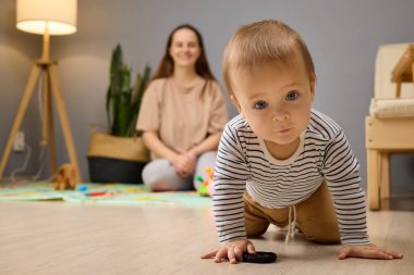 Küçük bir çocuk yerdeki küçük bir oyuncağa doğru sürünüyor. Bir anne yakınlardaki bir sandalyeden sevgi dolu bir şekilde izler, iyi aydınlatılmış bir oturma odasında sıcak ve besleyici bir atmosfer yaratır..