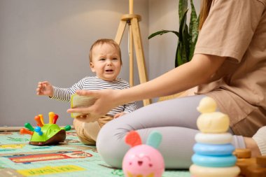 Bir bebek, çeşitli oyuncaklarla çevrili renkli bir oyun hasırının üzerinde oturur. Bir yetişkin oyuncakla uzanıp besleyici bir ortam yaratırken çocuk üzgün görünür..