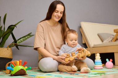 Bir anne ve yeni yürümeye başlayan çocuğu, rahat oturma odalarında renkli bir oyun hasırının üzerinde ahşap oyuncaklarla oynuyorlar. Sıcak atmosfer yaratıcılığı ve bağlanmayı teşvik eder..