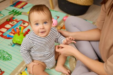 Bir bebek canlı bir oyun hasırının üzerinde oturur, oyuncakları keşfederken gülümser. Bakıcı, çocukla rahat bir ortamda kibarca etkileşime girer ve oyun zamanı neşesini arttırır..
